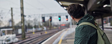 Young man with a backpack on a railway platform gazing down empty tracks on an overcast day.