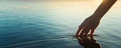 Hand gently touching calm ocean water at golden hour, creating soft ripples with a sunset horizon.