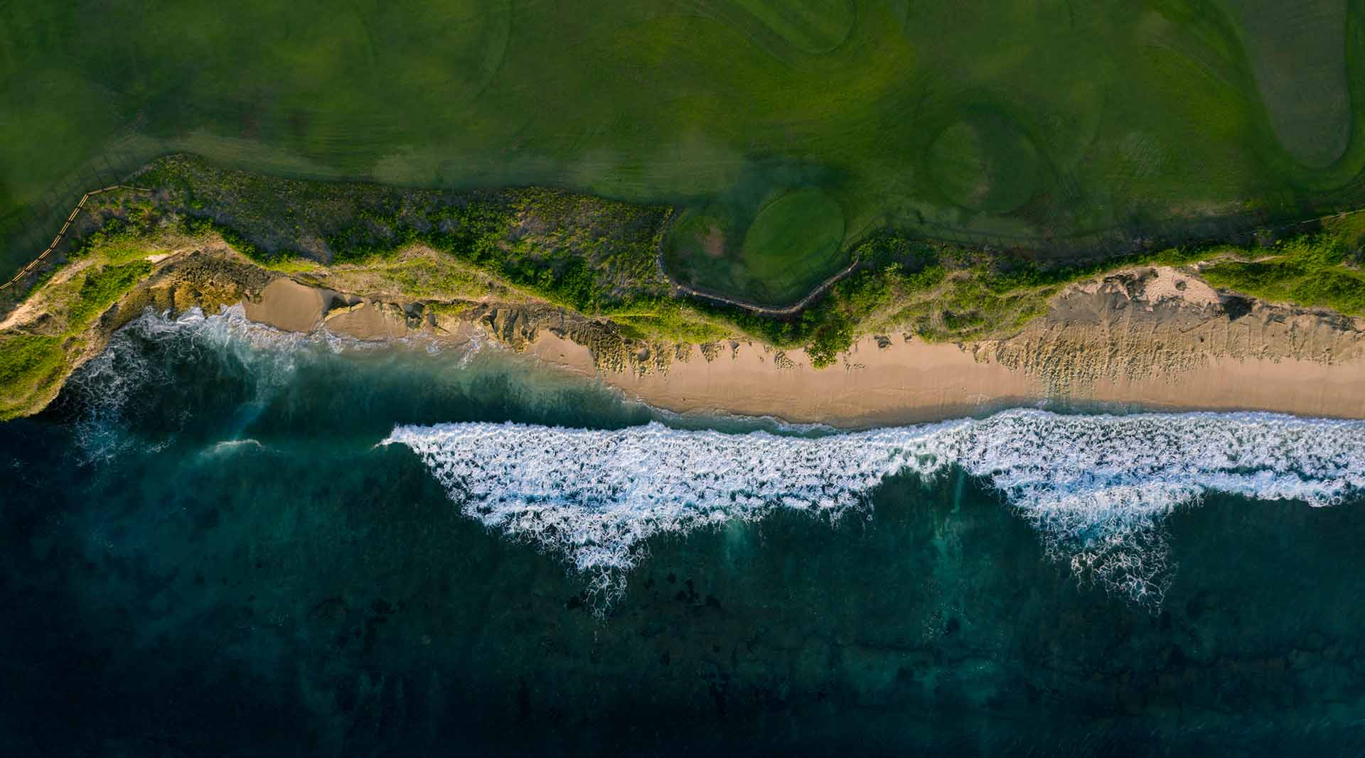 Aerial view of a rugged coastline with green cliffs, sandy beach, and ocean waves breaking along the shore