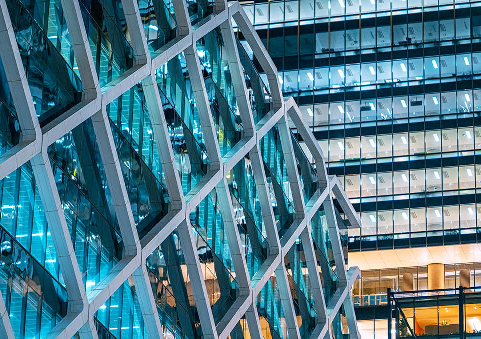 Night view of a modern glass office building with teal-lit diamond facade. Used in the private capital call series.