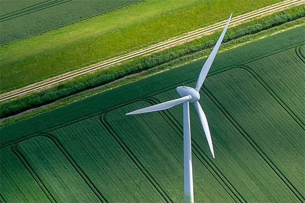 Aerial view of a white wind turbine with three blades over lush green agricultural fields with visible crop rows and a dirt path