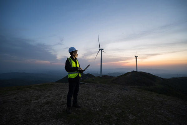 Data center energy article: Technician with laptop inspecting wind turbines on a hillside at dusk, clean energy concept.