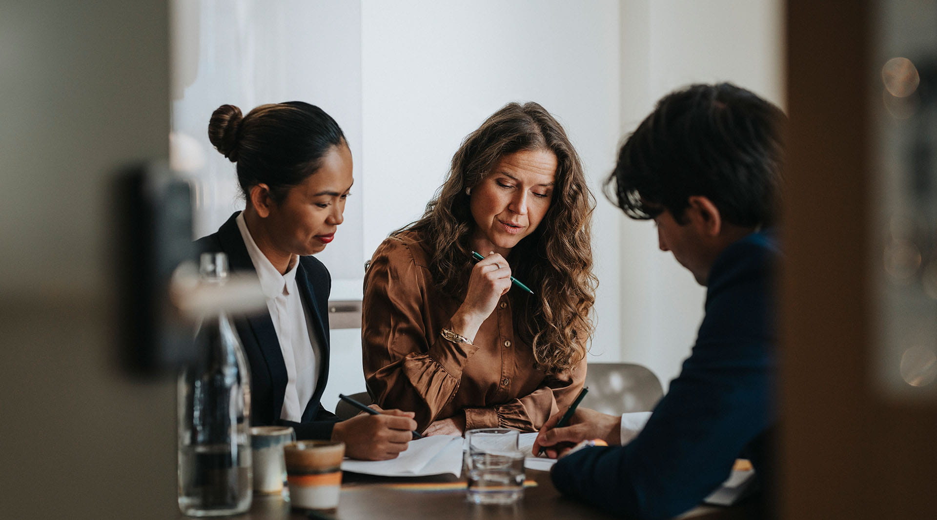 Three women reviewing documents together at a dark wooden conference table in a bright meeting room