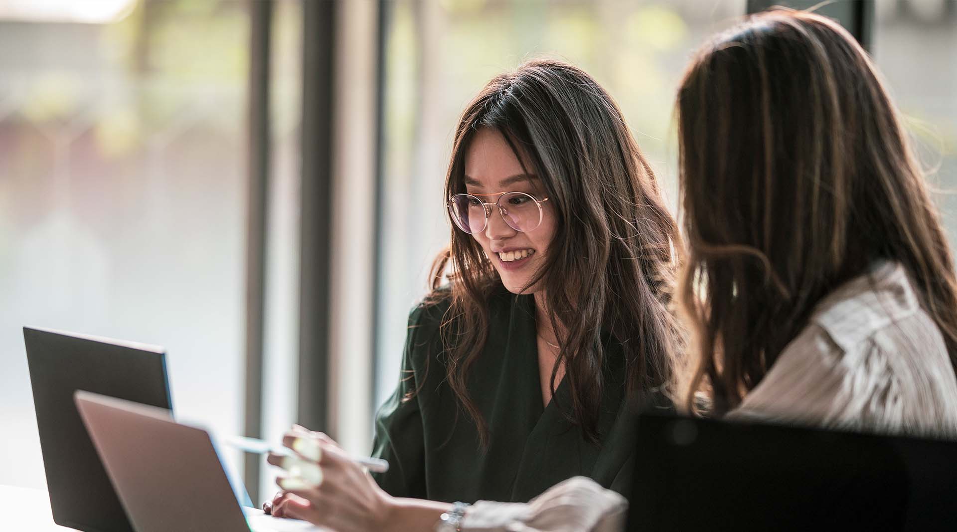 Two young women collaborating over a laptop in a modern office space with natural light and greenery