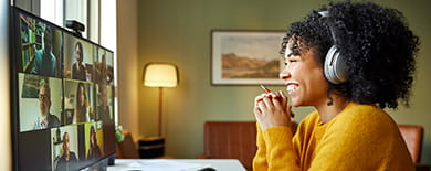 Smiling woman wearing headphones on a video conference call at a home office desk in warm lighting.