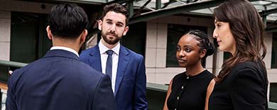 Four young professionals in business attire chatting outside a modern corporate building under an overcast sky.