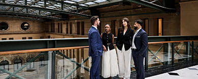 Four graduates standing on a mezzanine walkway in a heritage sandstone building with arched windows and marble floors.