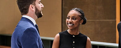 A young man and woman in professional attire smiling and talking in a modern corporate atrium with bronze wall panels.