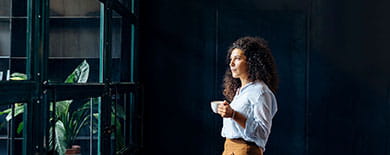 Young woman with curly hair holding a coffee cup and gazing thoughtfully in a stylish office with indoor plants.