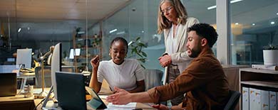  Three colleagues collaborating over documents and laptops at a desk in a modern open-plan office in the evening.