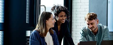 Three young professionals smiling and collaborating around a laptop in a bright office with exposed brick walls.