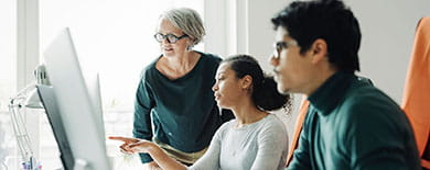 A senior colleague guiding two young interns as they review work on a desktop monitor in a bright, modern office.