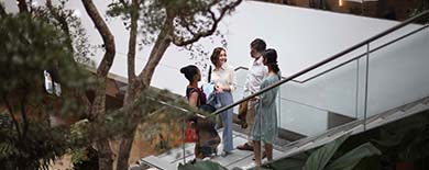Four young professionals chatting on a walkway inside a modern atrium building with indoor trees and greenery