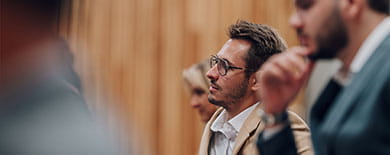 Young man in glasses and a tan blazer listening attentively during a professional seminar or conference