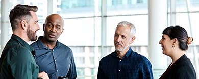 Four colleagues having a casual standing conversation in a bright office lobby with floor-to-ceiling windows