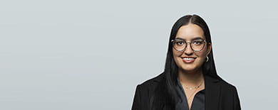 Professional headshot of a smiling young woman with long dark hair, glasses, and a black blazer