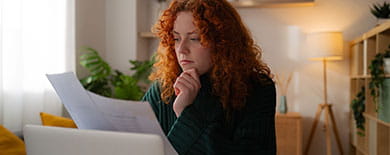 Young woman with curly red hair reading a document at her desk in a cozy home office with plants and warm lighting