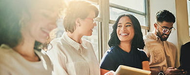 Four young professionals smiling together near a sunlit office window, one holding a tablet