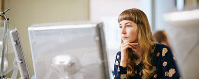 Young woman with bangs resting her chin on her hand while looking thoughtfully at a computer screen in an office