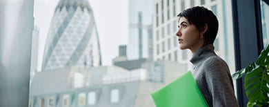 Young woman holding a green folder gazing out an office window at London's skyline including the Gherkin building