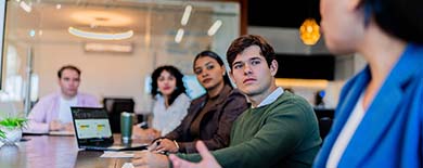 Five young professionals in a meeting around a conference table with a laptop, in a modern room with glass walls