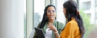 Two women having an animated conversation in a bright office with floor-to-ceiling windows and a city view