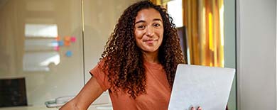 Young woman with curly brown hair holding a laptop and smiling confidently in a modern office with glass walls