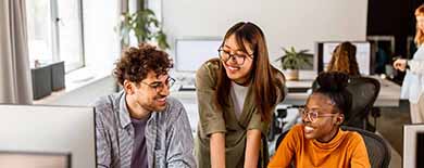Three young professionals smiling and collaborating around a laptop in a bright open-plan office with plants