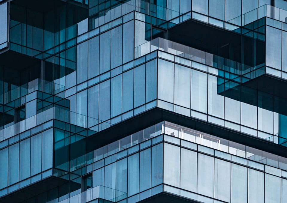 Modern glass high-rise building facade with geometric blue-teal panels, protruding balconies, and rectangular grid patterns