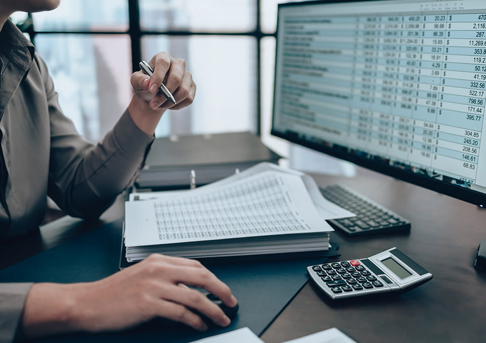 Employee reviewing a spreadsheet at a desk with a calculator, binder and paperwork. 