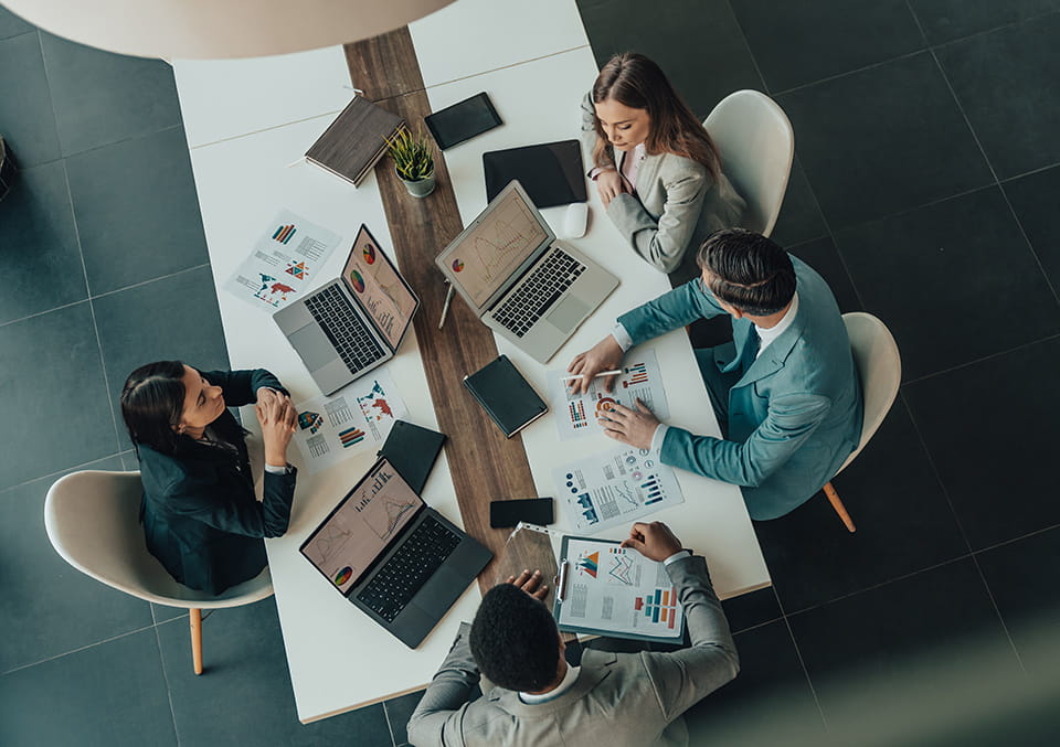 Four business professionals reviewing charts on laptops, a tablet and printed reports around a conference table.