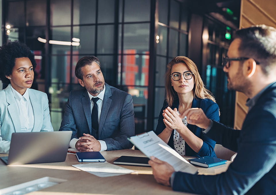 Four professionals discuss a contract in a modern office conference room with glass walls and warm lighting