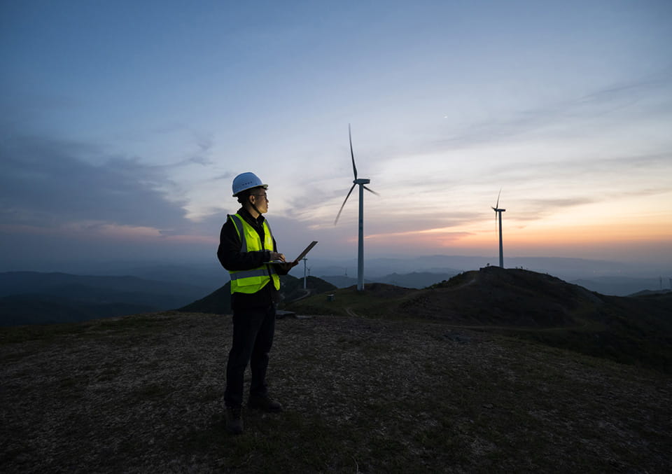 Data center energy article: Technician with laptop inspecting wind turbines on a hillside at dusk, clean energy concept.