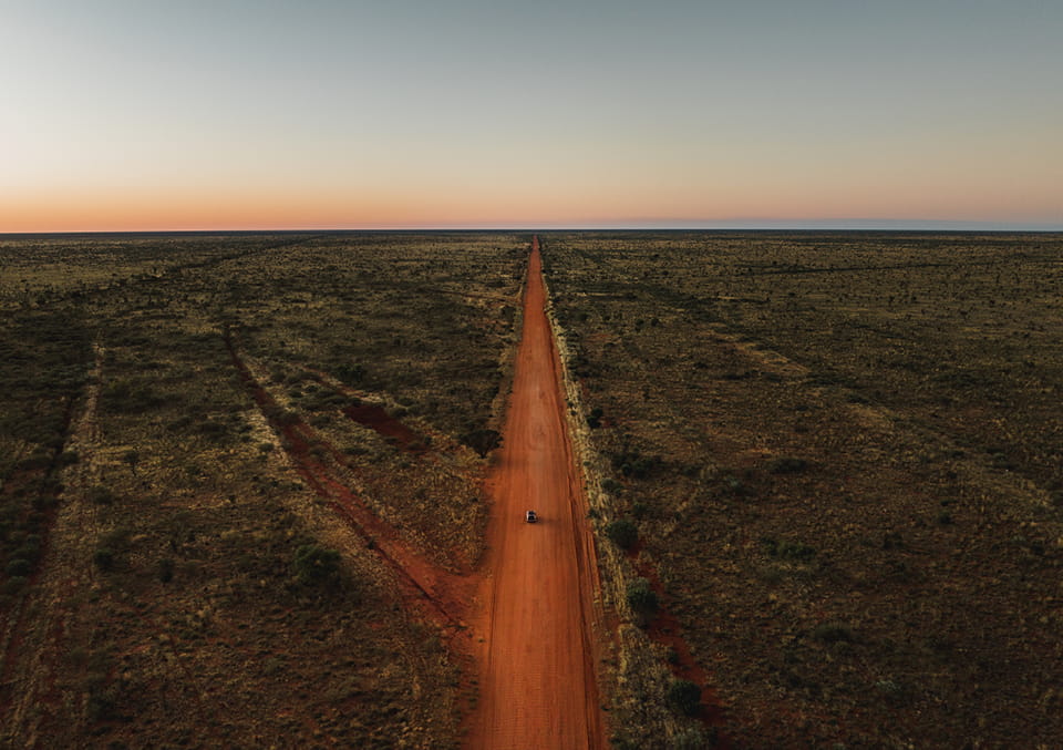 Aerial view of a straight red dirt road with a single vehicle, running through flat outback scrub towards a sunset horizon.