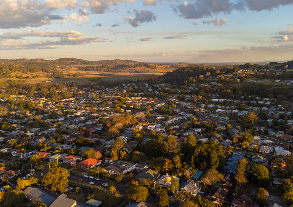 Aerial view of a small Australian town with mixed rooftops and mature trees, and rolling green and golden hills at golden hour.