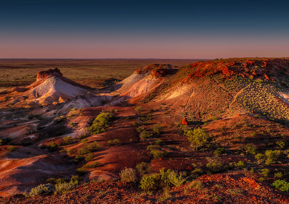 Eroded outback mesas with red, white and ochre rock bands above a vast plain at twilight.