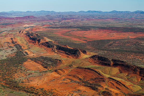 Aerial view of red, iron-rich outback ridges and escarpments with sparse scrub, and distant blue ranges beyond.
