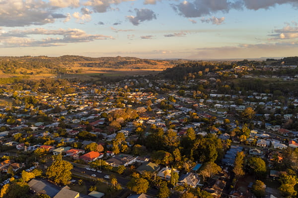 Aerial view of a small Australian town with mixed rooftops and mature trees, and rolling green and golden hills at golden hour.
