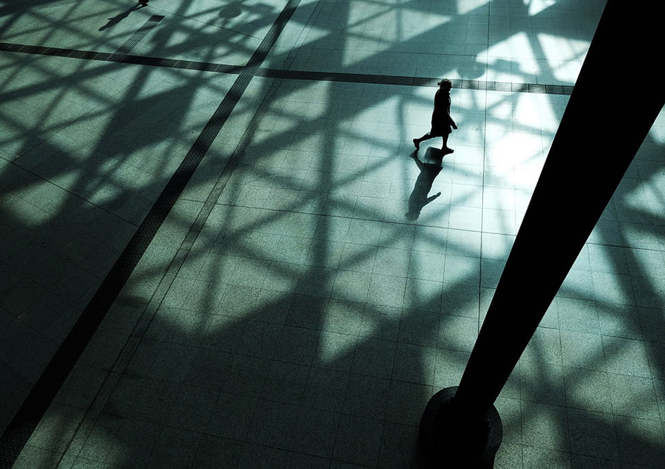 Aerial view of a lone figure walking through a modern atrium with geometric shadows cast by a glass and steel skylight