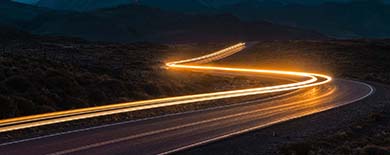 Long-exposure photograph of light trails from vehicles on a winding S-curve road at dusk with mountains in the background.