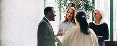 Four professionals smiling and conversing in a bright modern office with green-framed windows and indoor plants.