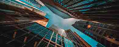 Dramatic upward view from street level of converging glass skyscrapers framing a narrow patch of blue sky.