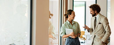 Two professionals in conversation in a modern office - a woman holding a tablet and a man in a tan suit gesturing.