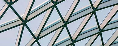 Upward view of a modern geometric glass and green steel roof structure against a pale blue sky