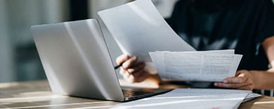 Woman reviewing printed documents at a wooden desk beside an open laptop in a sunlit office