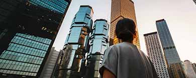 Woman with hair in a bun gazing up at modern Hong Kong skyscrapers against a golden sunset sky.