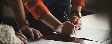 Two people signing legal documents at a dark wooden table, hands and papers in warm dramatic lighting.