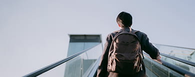 Person in a gray coat riding a glass-paneled escalator upward against a pale overcast sky.