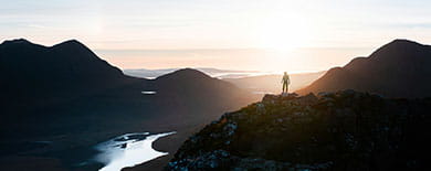 Silhouetted person standing on a mountain ridge overlooking a valley and river at golden sunrise.