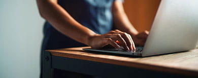 Person typing on a silver laptop at a wooden standing desk in soft warm lighting.